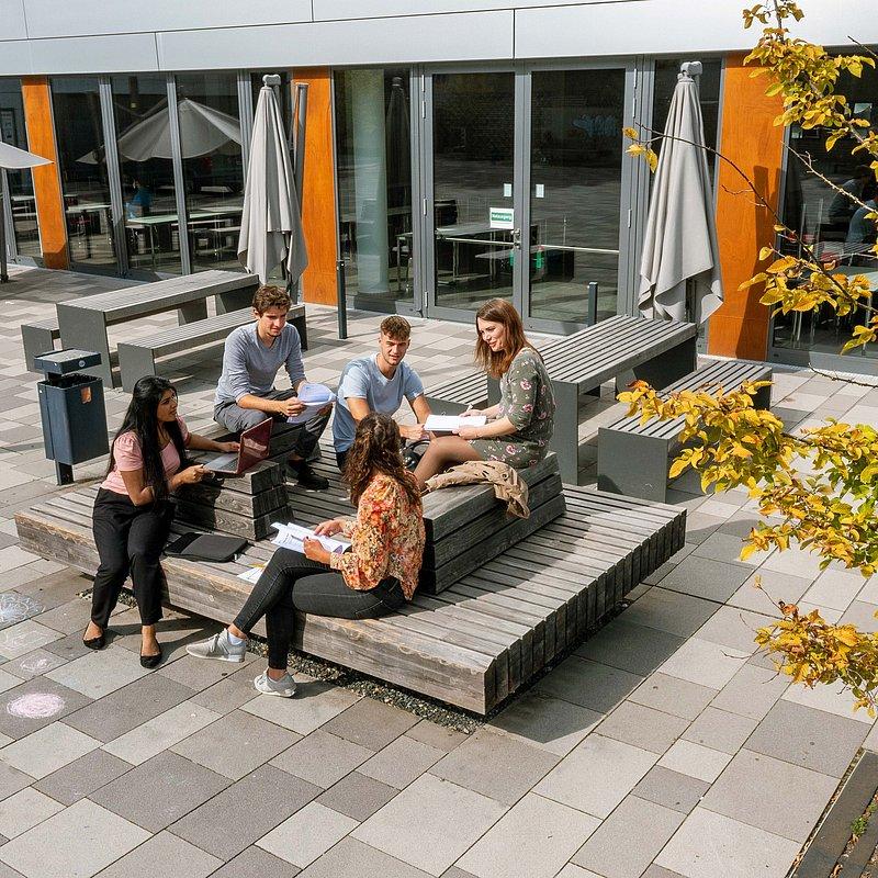 Gruppe von fünf Personen sitzt draußen auf Holzsitzen und lernt gemeinsam.