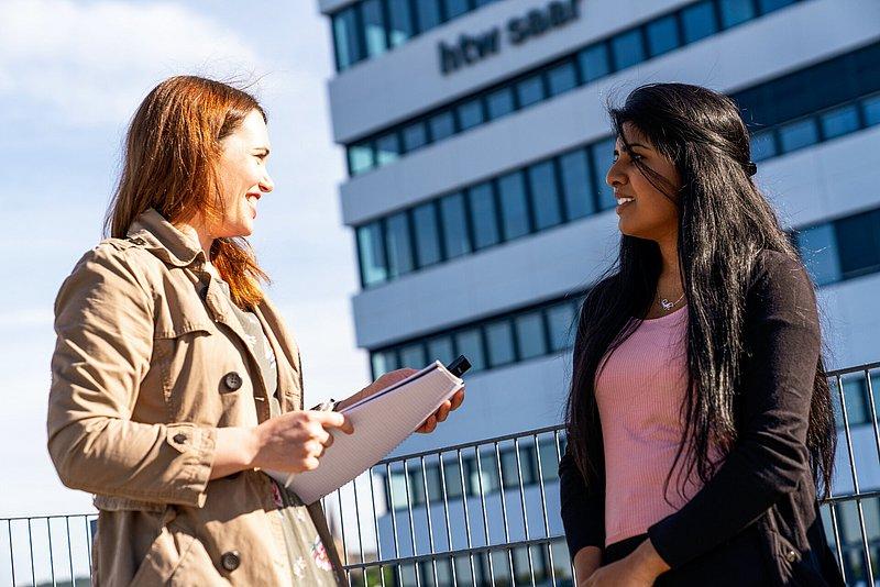 Zwei junge Frauen sprechen draußen vor einem Hochschulgebäude.