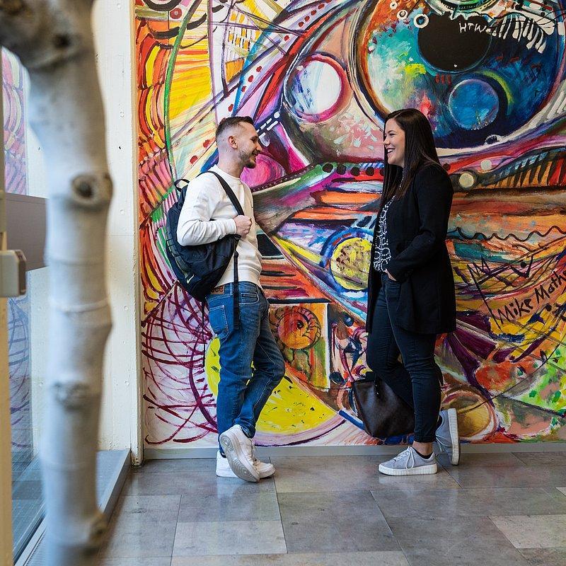 Two people talking in front of a colorful mural.