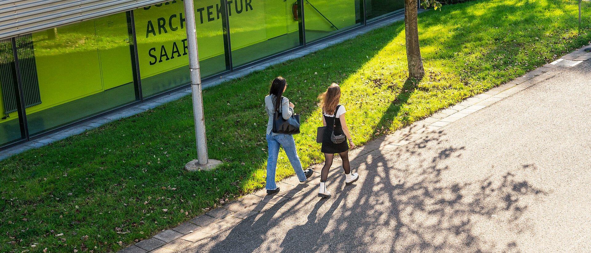 Zwei Personen gehen am Gebäude mit grüner Glasfront vorbei.