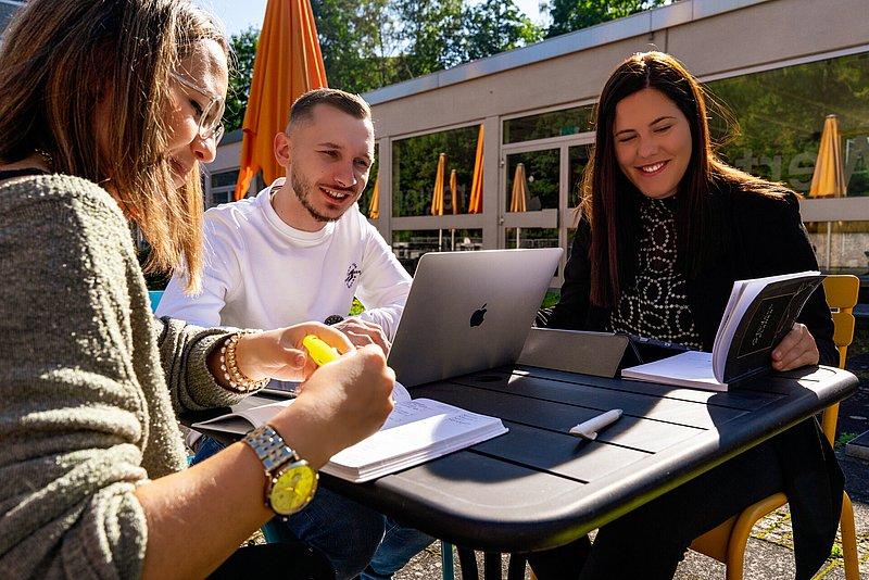 Three young adults sit outside at a table with a laptop, book, and notes.