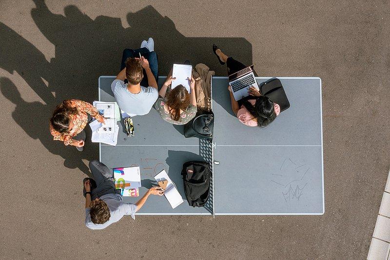 Fünf Personen arbeiten draußen auf einer Tischtennisplatte, von oben fotografiert.