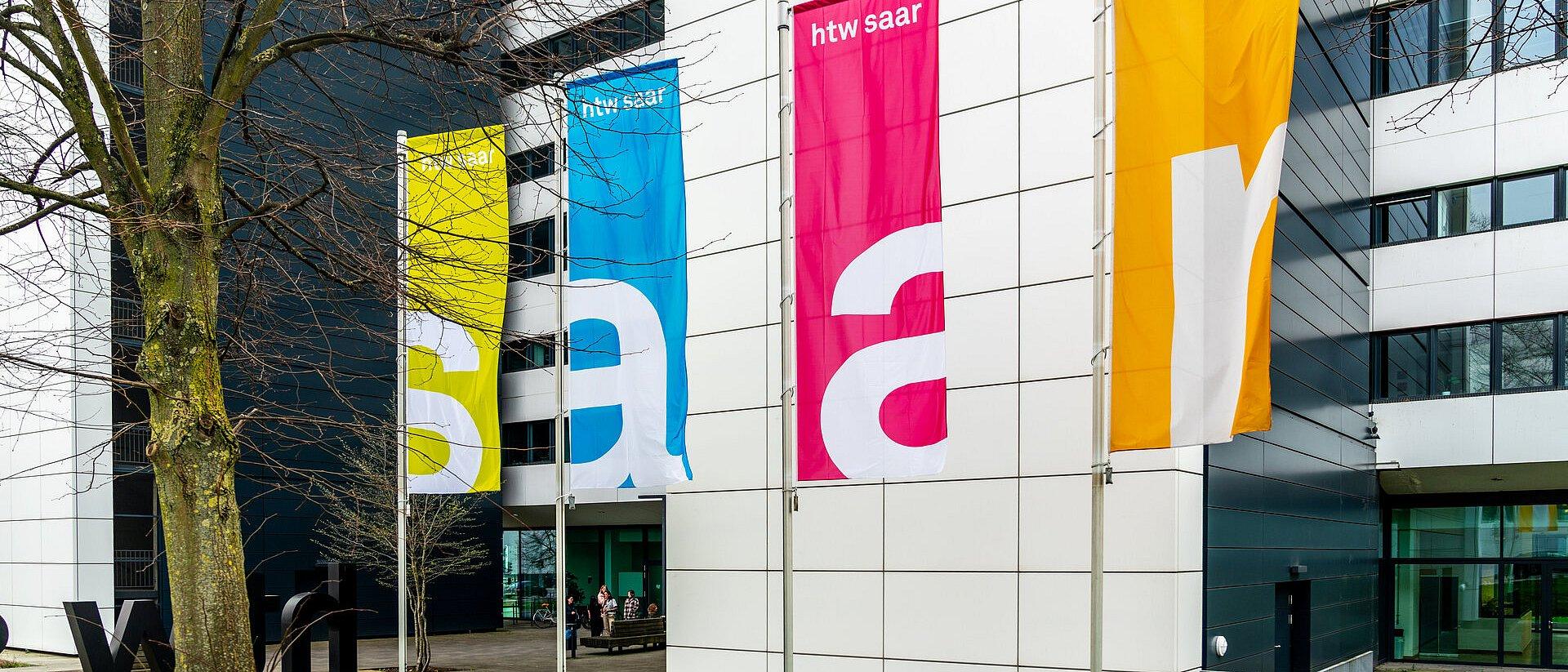 Four colorful flags with “htw saar” in front of a building.