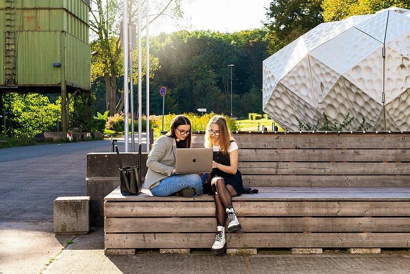 Zwei junge Frauen sitzen draußen auf Holzstufen und arbeiten an einem Laptop.