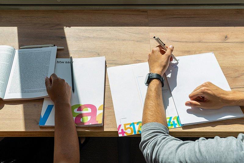 Zwei Personen arbeiten an Schreibtisch mit Büchern und Blättern in der Sonne.