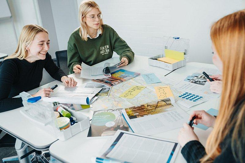 Drei junge Frauen blättern in Magazinen an einem Tisch mit Karten und Notizen.