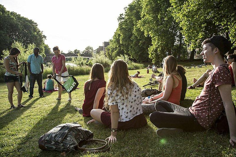 Mehrere Menschen sitzen und stehen auf einer sonnigen Wiese im Park, einige mit Taschen oder Fahrrädern; im Hintergrund ein Fluss und eine Brücke.