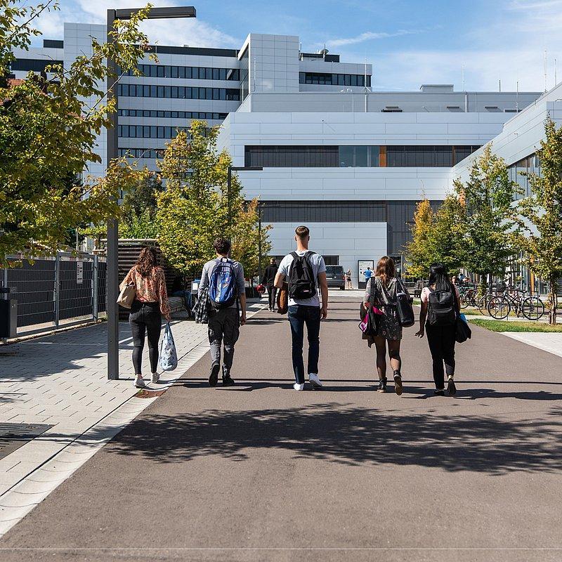 Five people with backpacks walking toward a modern university building.