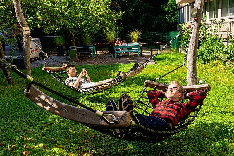 Two young men lie in hammocks in the green, one looking at his phone.