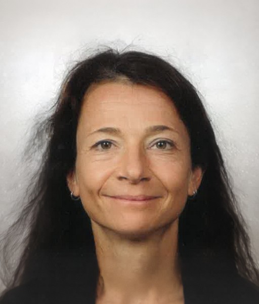 Porträtfoto einer Frau vor hellem Hintergrund.   Die Frau hat mittellange, dunkle Haare.   Sie hat einen hellen Teint und trägt kleine, runde Ohrringe.   Ihr Gesicht ist frontal aufgenommen, sie lächelt leicht mit geschlossenen Lippen.   Die Augen sind offen und blicken geradeaus.  
