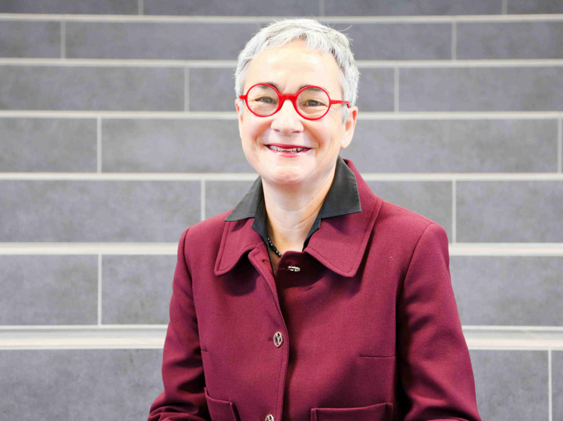 Das Foto zeigt Prof. Dr. Ulrike Zöller. Sie hat an der htw saar eine Professur für Theorie, Methodik und Empirie Sozialer Arbeit. Abgebildet ist sie freundlich lächelnde Frau mit kurzem grauem Haar.   Sie trägt eine runde, auffällige rote Brille und hat einen hellen, rosigen Hautton.   Die Lippen sind mit einem rötlichen Lippenstift geschminkt, die Augenbrauen sind kurz und eher hell.   Sie trägt einen dunkelroten, fast bordeauxfarbenen Mantel oder Blazer mit großem Kragen und Knöpfen vorne. Der Hintergrund besteht aus grauen, waagerecht verlaufenden Stufen oder Fliesen, wodurch die Person klar im Vordergrund steht.