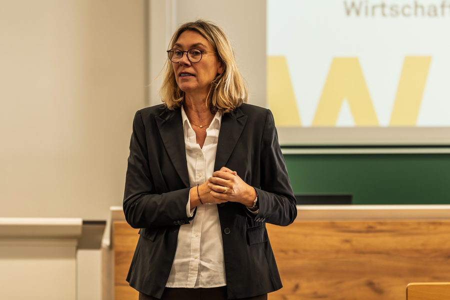 Das Bild zeigt Prof. Dr. Petra Garnjost, Professur für Allgemeine Betriebswirtschaftslehre an der htw saar. Sie steht in Businesskleidung vor einer Präsentationsfläche.  Sie hat schulterlanges, blondes Haar, helle Haut und trägt eine runde Brille mit dunklem Rahmen.   Sie trägt ein weißes Hemd, darüber ein dunkelgraues oder schwarzes Sakko.   Ihre Hände sind vor dem Körper verschränkt, die Finger locker ineinander gelegt.  